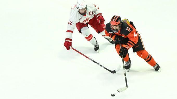Two hockey players, one in orange and the other red, compete for a puck.