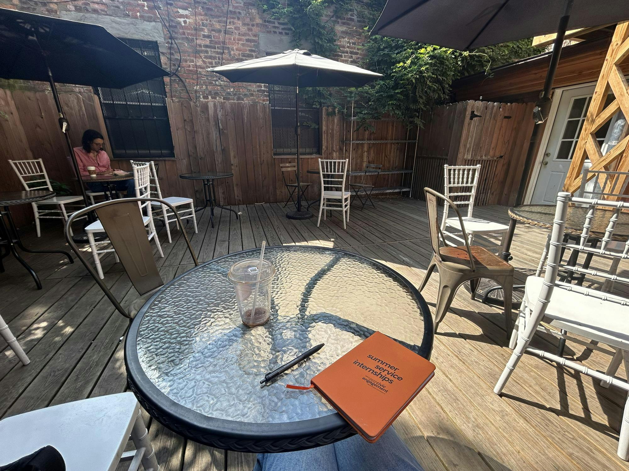 A circular glass table with a small orange notebook, pen, and empty plastic coffee cup, surrounded by other chairs and tables strewn across wooden patio floorboards. 