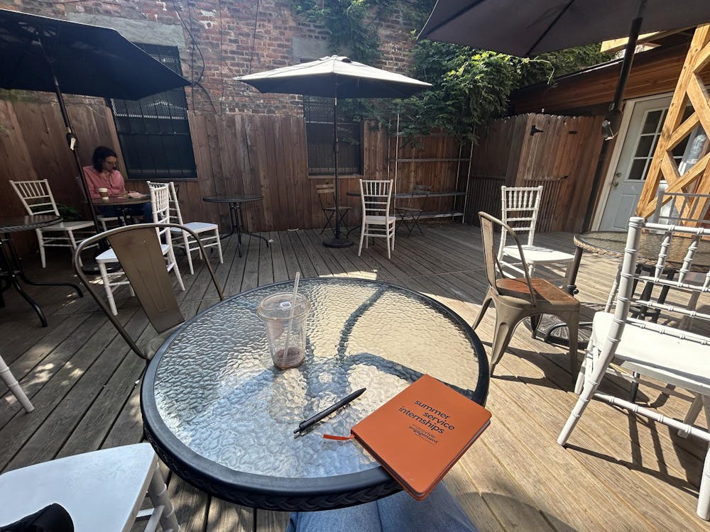 A circular glass table with a small orange notebook, pen, and empty plastic coffee cup, surrounded by other chairs and tables strewn across wooden patio floorboards. 
