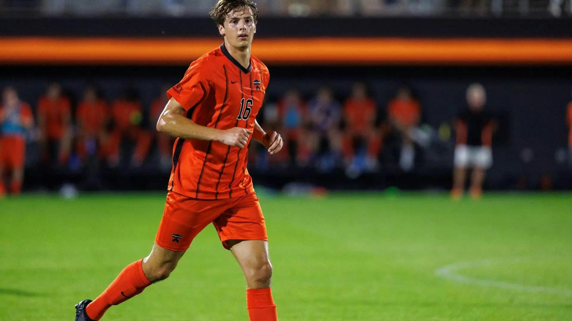 A man wearing orange shorts and an orange jersey jogging on a soccer field.