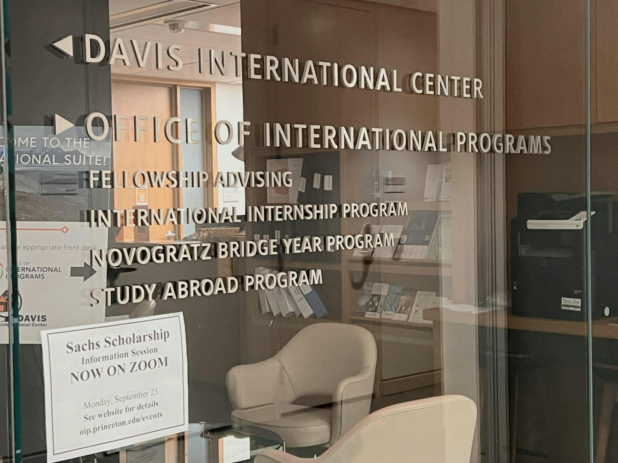 A glass door can be seen, with text on it that reads, “Davis International Center,” “Office of International Programs,” “Fellowship Advising,” “International Internship Program,” “Novogratz Bridge Year Program,” and “Study Abroad Program.” White armchairs and wooden shelving can also be seen through the glass doors.