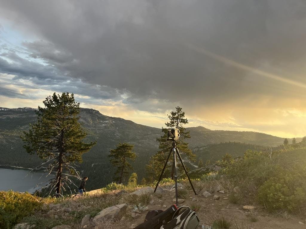 Large black camera on a black tripod is set up in front of gray and green mountains and a blue lake.