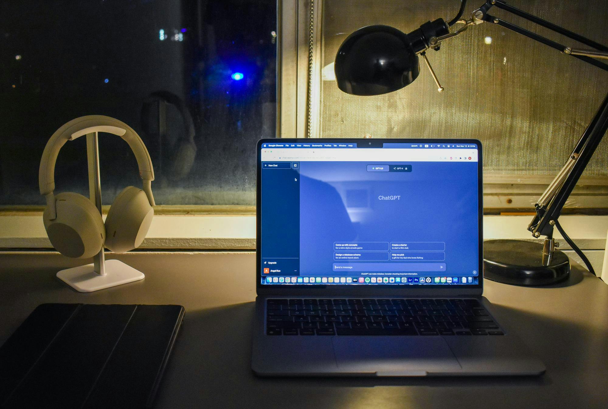 A computer sits on a student's desk next to a lamp and pair of headphones. Its blue light contrasts with the night of the window behind it. The computer screen has ChatGPT open.