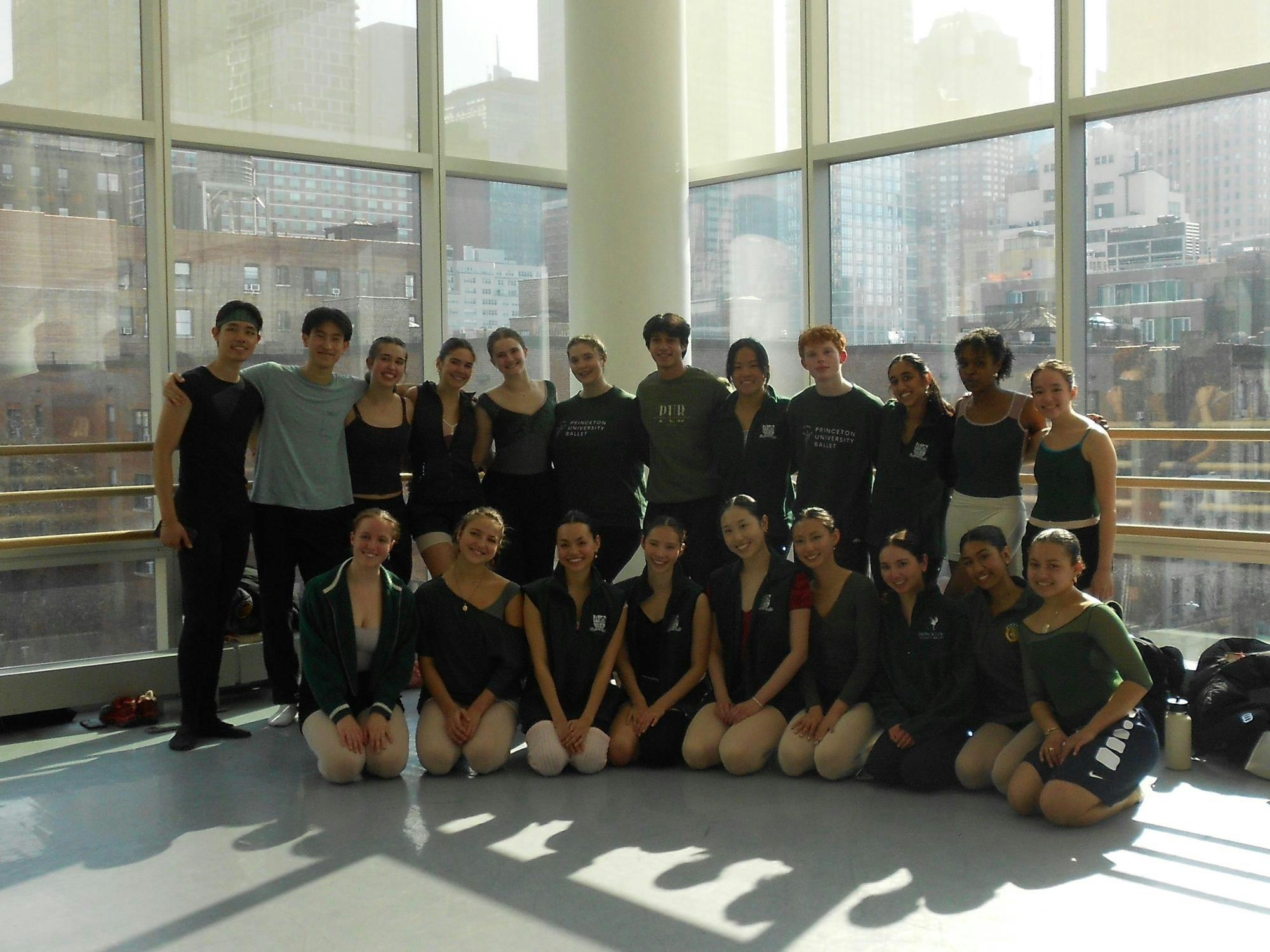 Dancers in mostly black outfits sit and stand in front of a large window through which one can see New York City buildings.