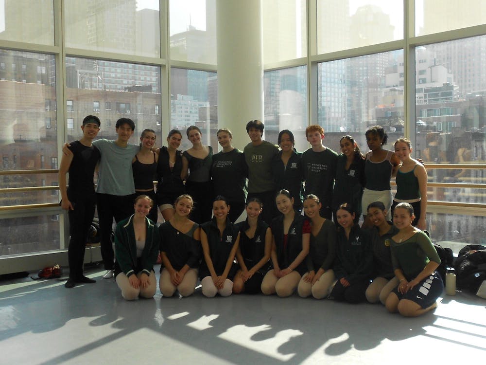 Dancers in mostly black outfits sit and stand in front of a large window through which one can see New York City buildings.