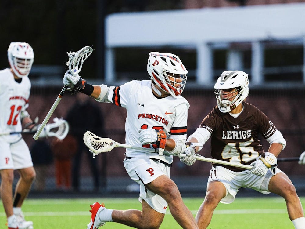 Man in a white Princeton jersey cradles ball in lacrosse stick as a defender in brown pokes under his arm with his own stick.