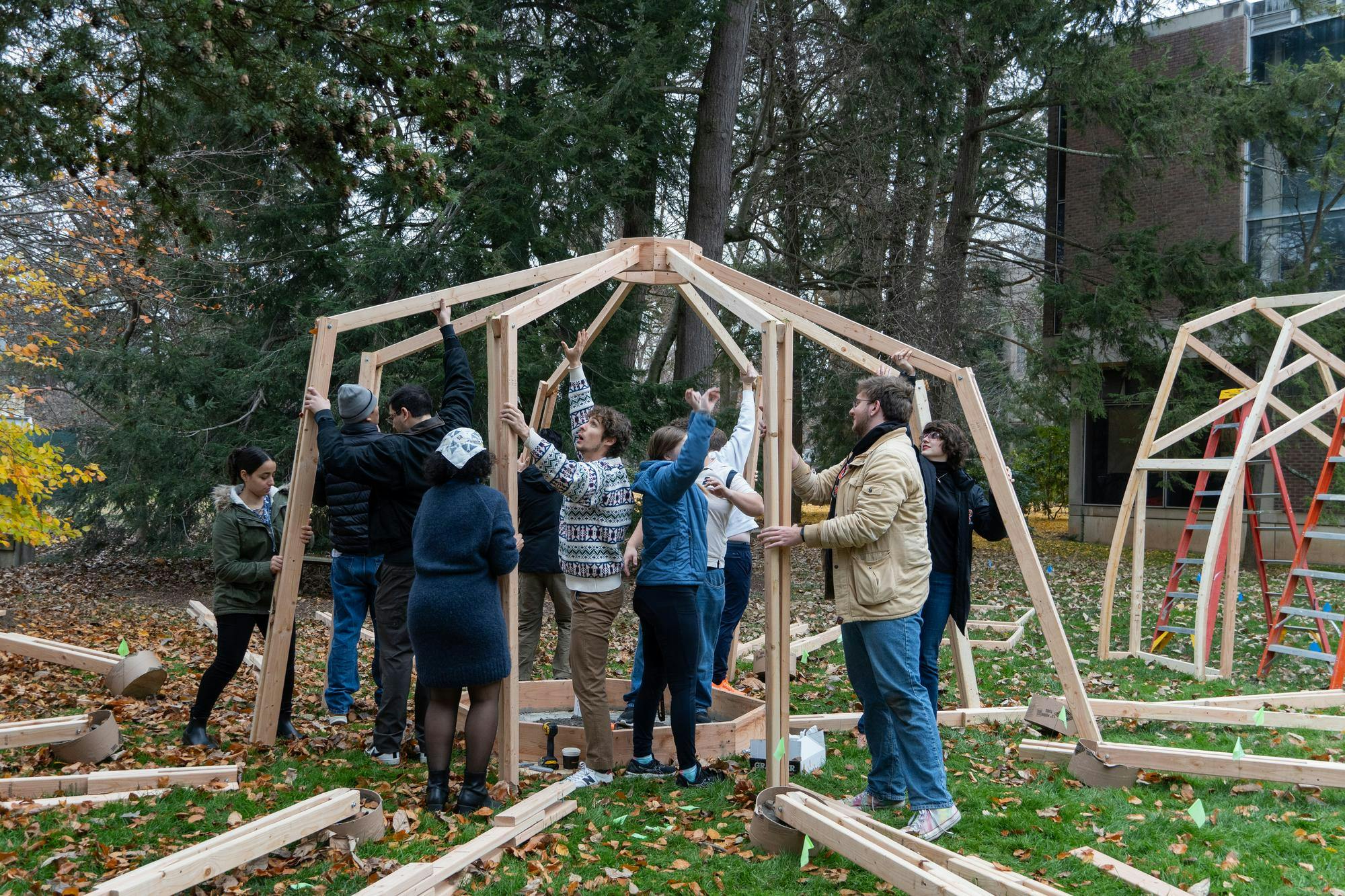 Many students in sweaters and jackets stand outside compiling lightly colored wood pieces into a circle.