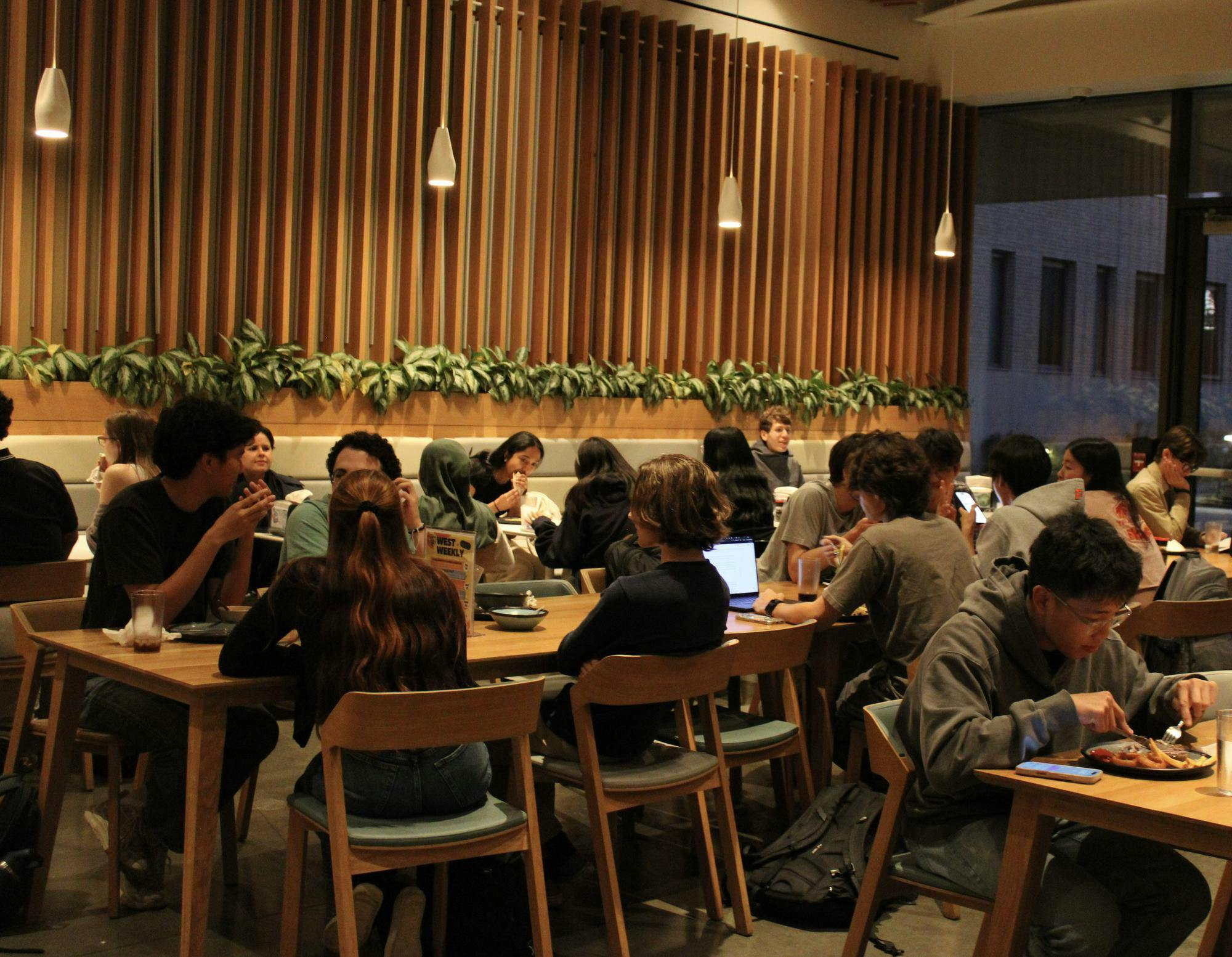 College students sit around rows of long wooden tables eating dinner, with lights and a planter in the background.