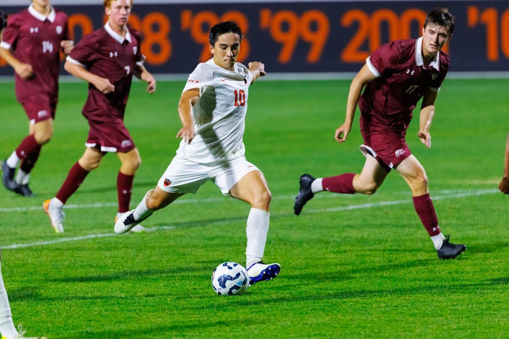 A man wearing white shorts and a white and orange jersey marked with the number 10 is kicking a soccer ball on a grass field.