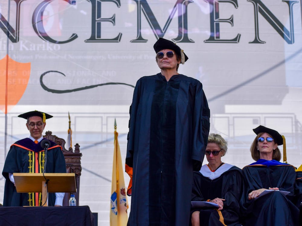 Woman in gown with flat hat with blue vestaments standing in front of four other people in gowns in front of a white background which has the letters “NCEMEN”. One of the people in the background has an orange lining, is bispectacled and is at a podium.