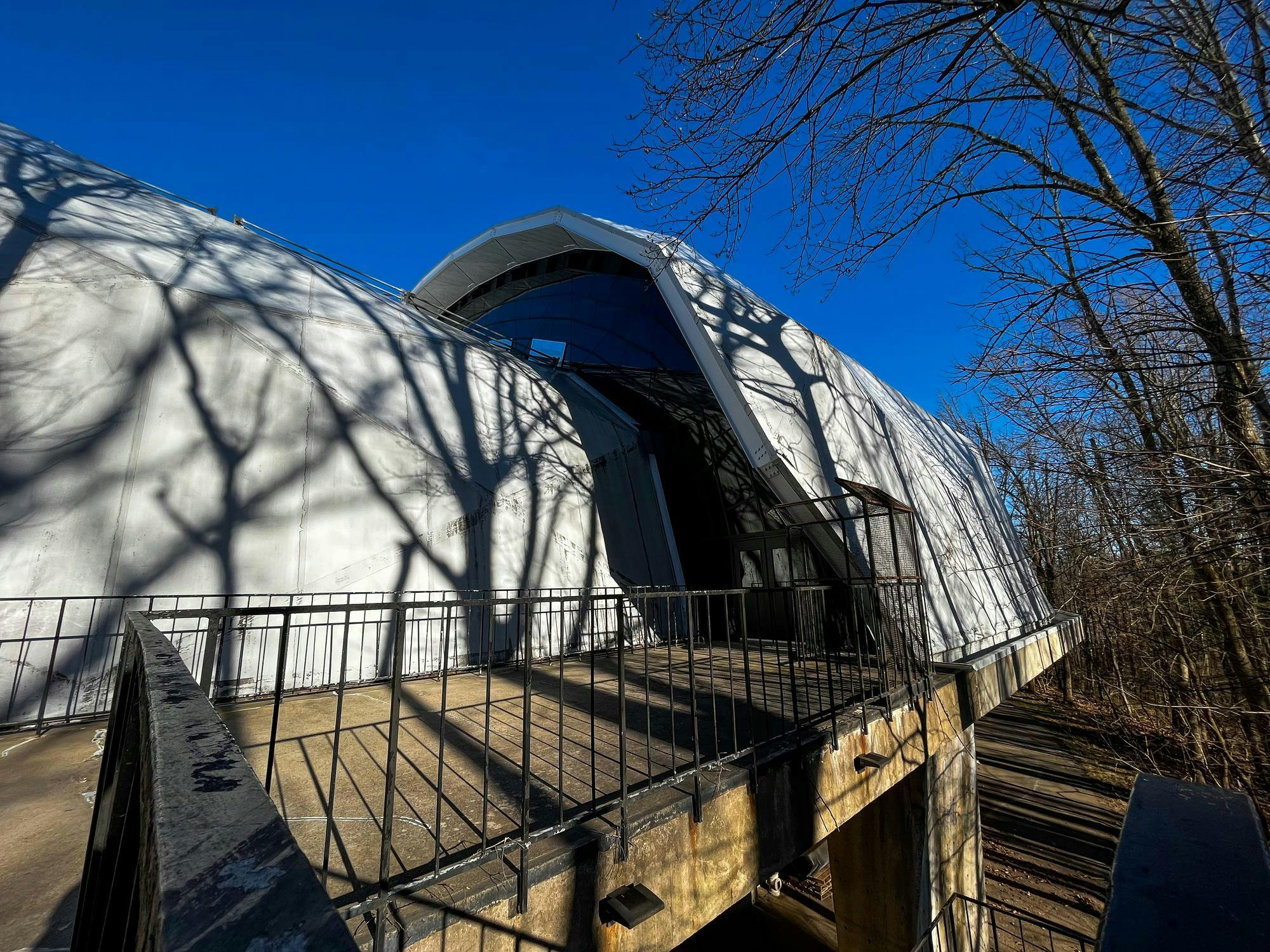 shadows of trees in the winter reflect on a white arched building with a balcony with a rusted railing.  It is a sunny day with a blue sky.