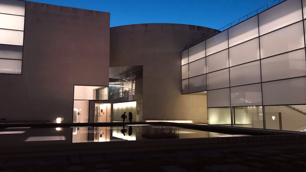 A large, vaguely brutalist building surrounds a reflecting pool. The sky is dark blue in nighttime. Bright lights shine from inside the large glass windows.