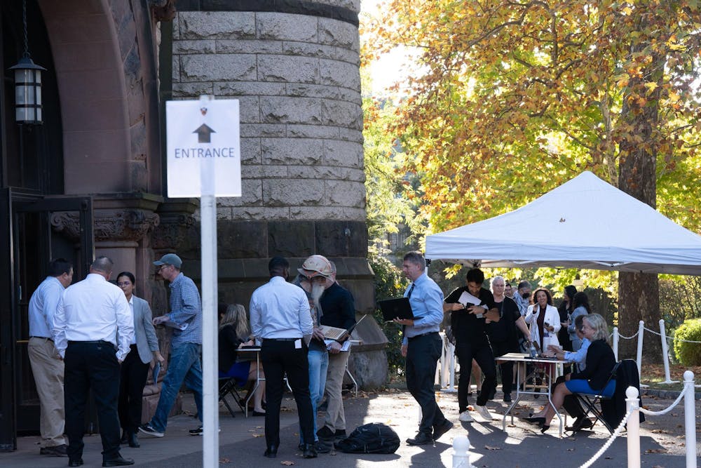 A group of people line up outside of a stone building. An entrance sign is in the foreground of the photo, and fall foliage rests in the background.