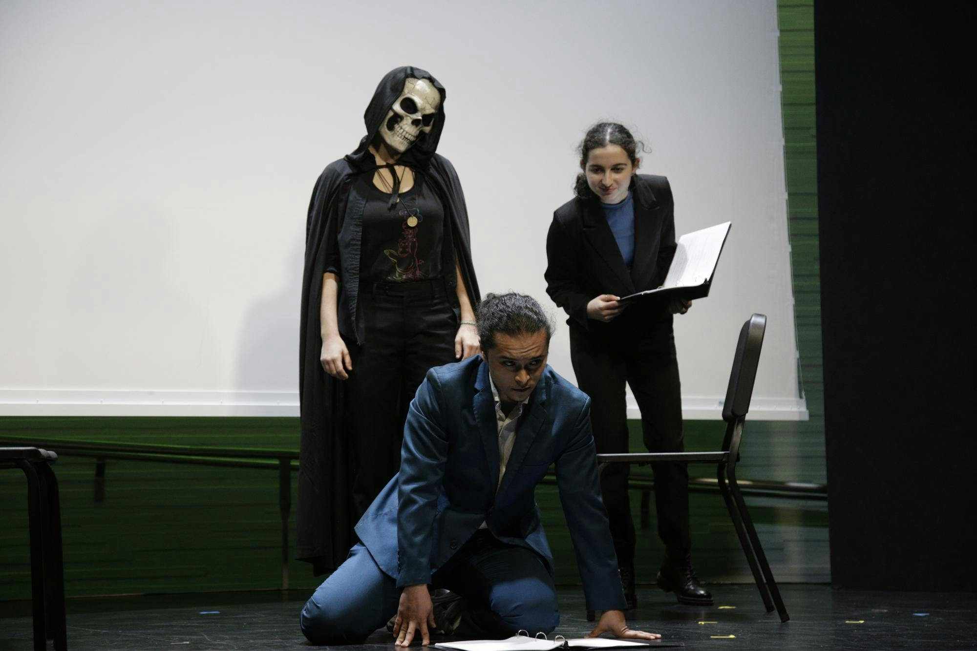 Person in dark, black cloak and skeleton mask and another person in a black suit watch on as a someone in a blue suit is on the ground.