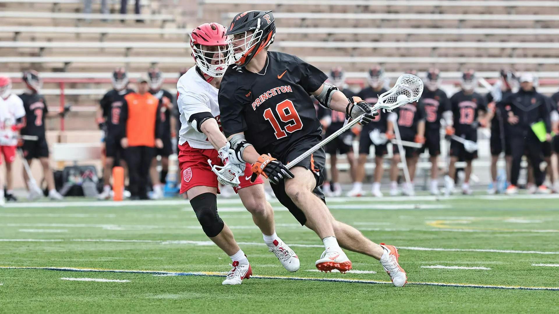 A man carrying a lacrosse stick with the ball in it while being guarded by his defender on a grass field. 
