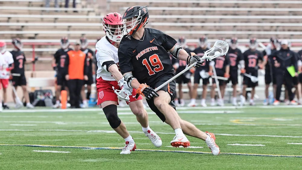 A man carrying a lacrosse stick with the ball in it while being guarded by his defender on a grass field. 
