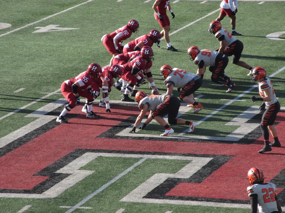 Football players in red and orange stand on a green turf field with a large H on it.