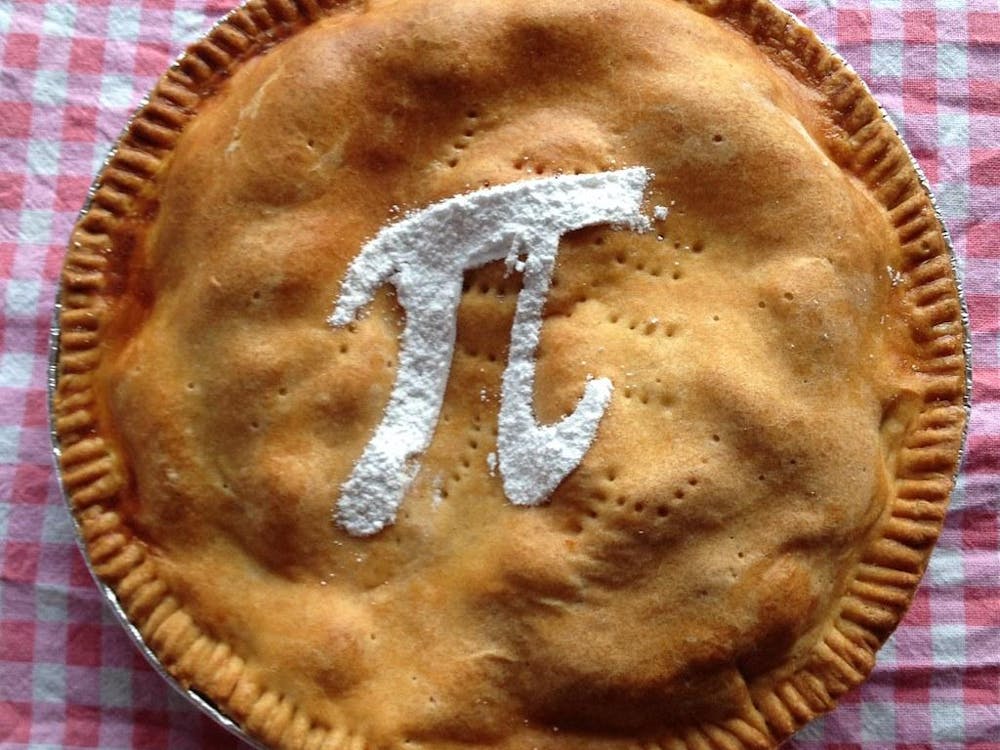 A golden circular pie with pinched crust on top of a red and white gingham background. In the middle of the pie, there is a white pi symbol that appears to be made of powdered sugar. 