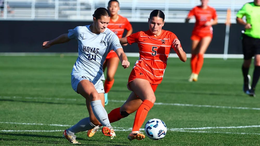 A woman wearing an orange jersey dribbling a ball with a defender wearing a white jersey chasing her down on a soccer field. 