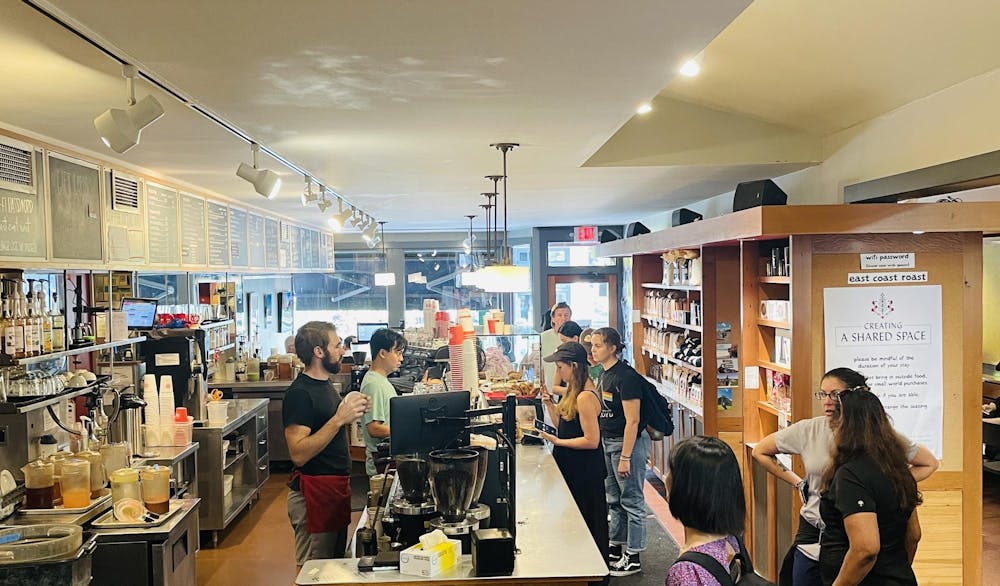 Customers line up to order coffee in front of a wooden shelf lined with coffee merchandise, while baristas prepare drinks.