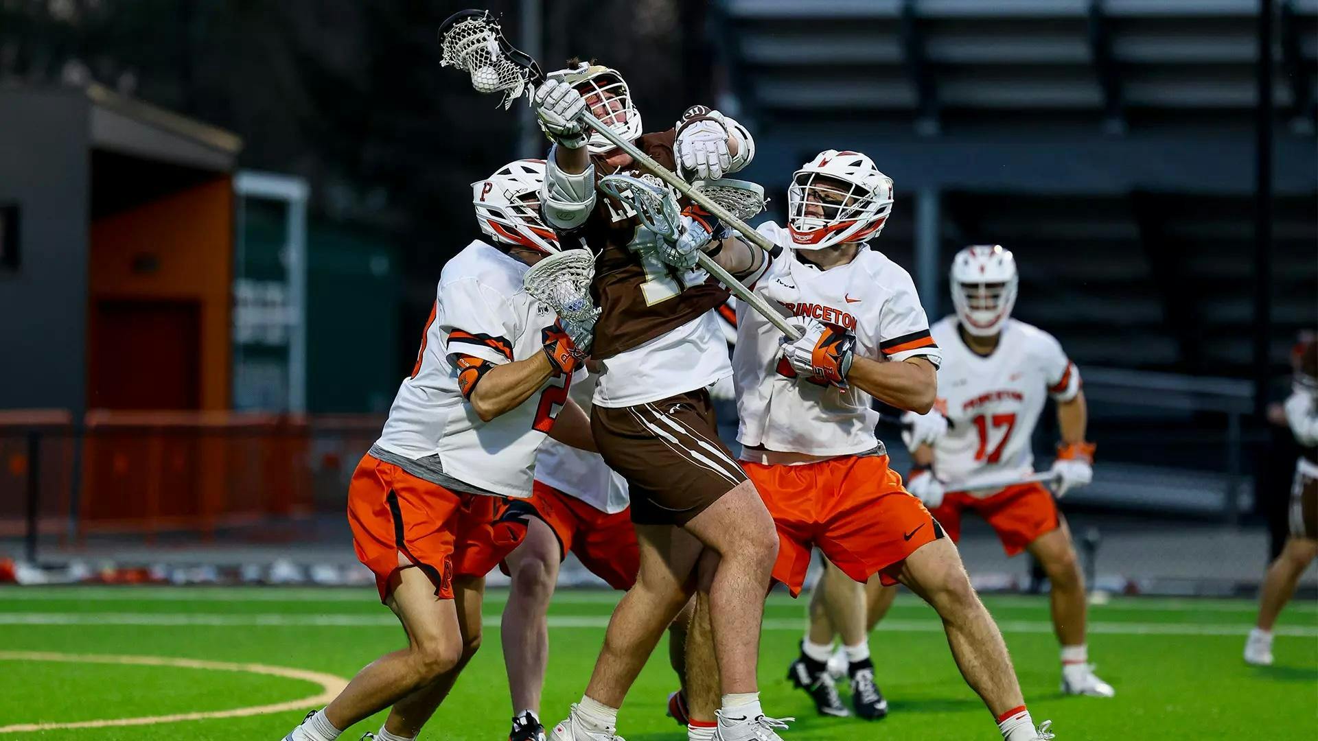 Two Princeton lacrosse players in white jerseys defending a player in a brown jersey.