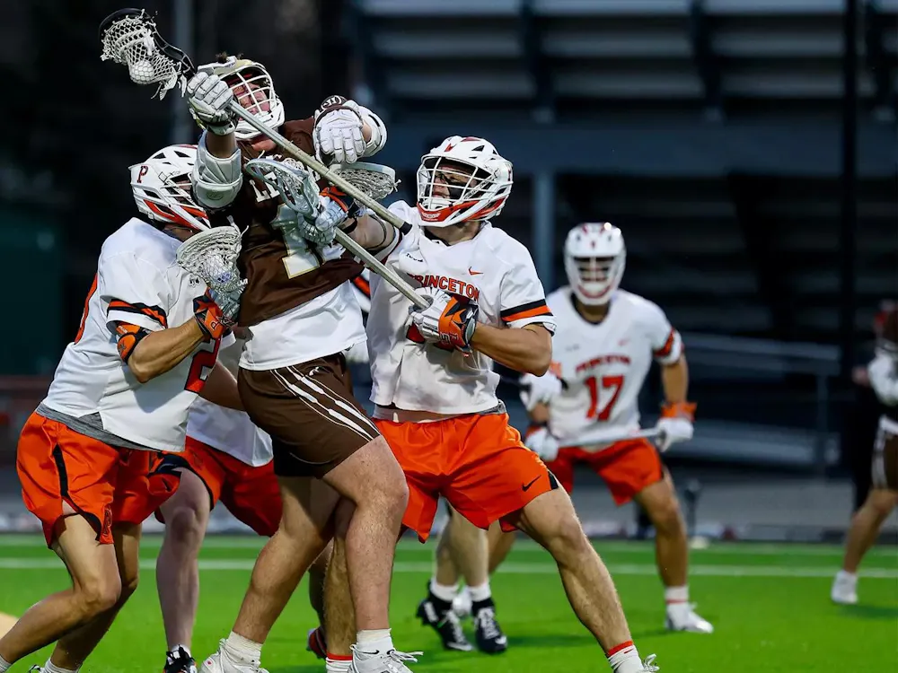 Two Princeton lacrosse players in white jerseys defending a player in a brown jersey.