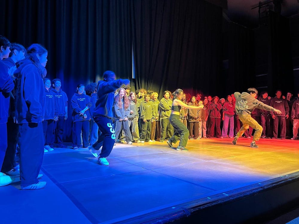 Dozens of students in dark outfits are in a half circle on a stage. In the middle of the circle, three dancers make a choreographed movement.