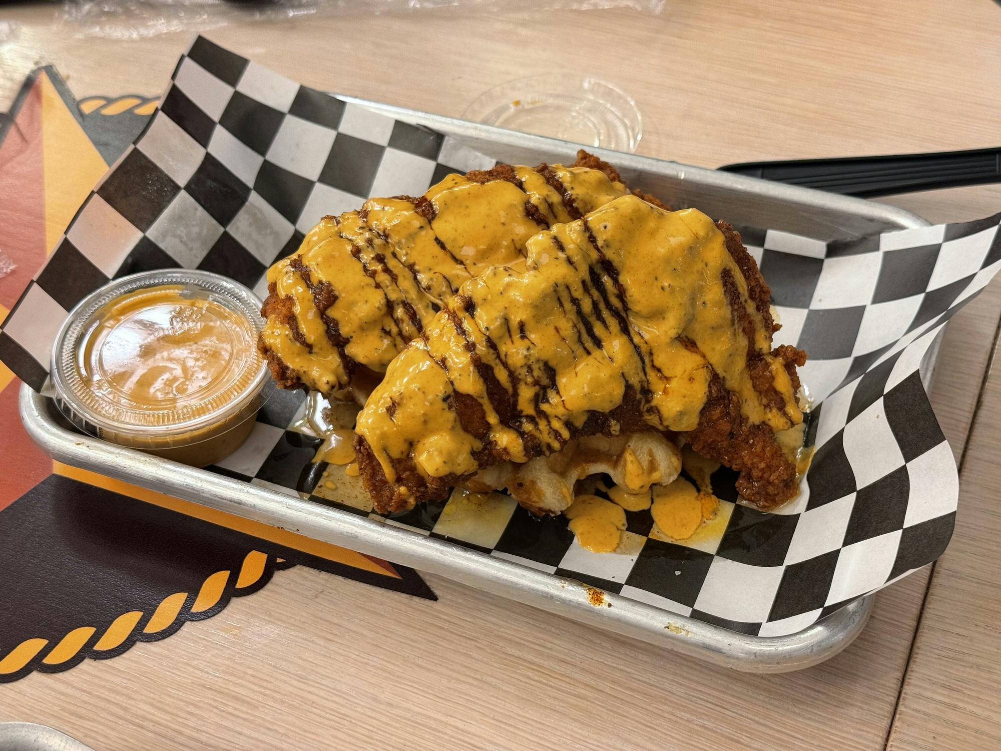 A small grey tray is lined with black and white checkered paper. There are two pieces of large, crispy, textured orange colored chicken. There is a small brown waffle that is slightly visible under the chicken. There is yellow sauce all over the chicken. There is also a small transparent container of yellow sauce in it.