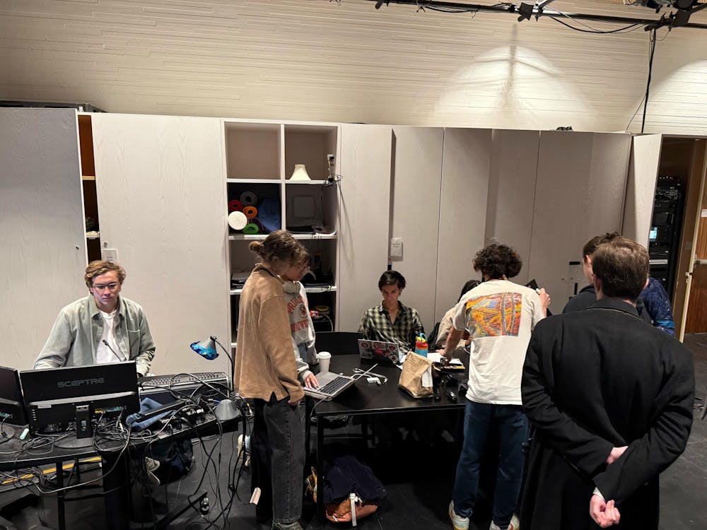 Several people are gathered around two tables, with some working on their computers, some collaborating and discussing, and a few observing the situation.  