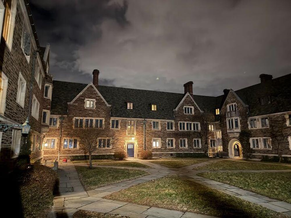 A brown building with a black roof has most windows dark. The sky is dark and cloudy, and the ground has grass and gray pathways.