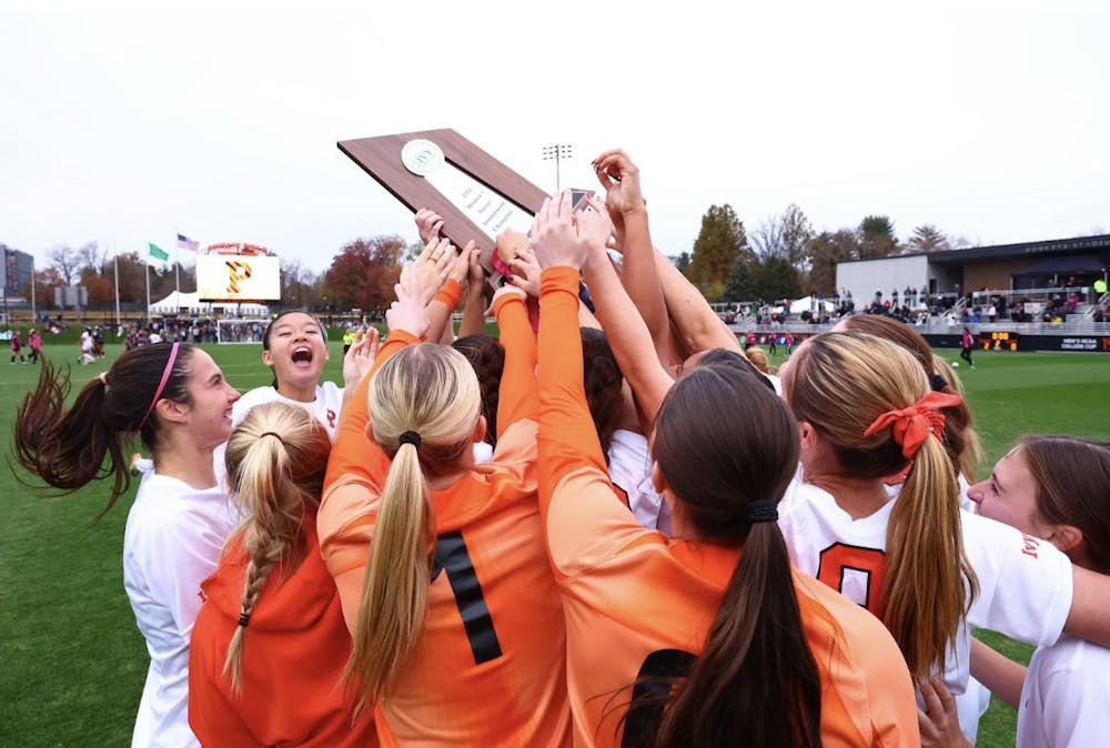 A women's soccer team celebrates a victory, raising a trophy over their huddle on the field.