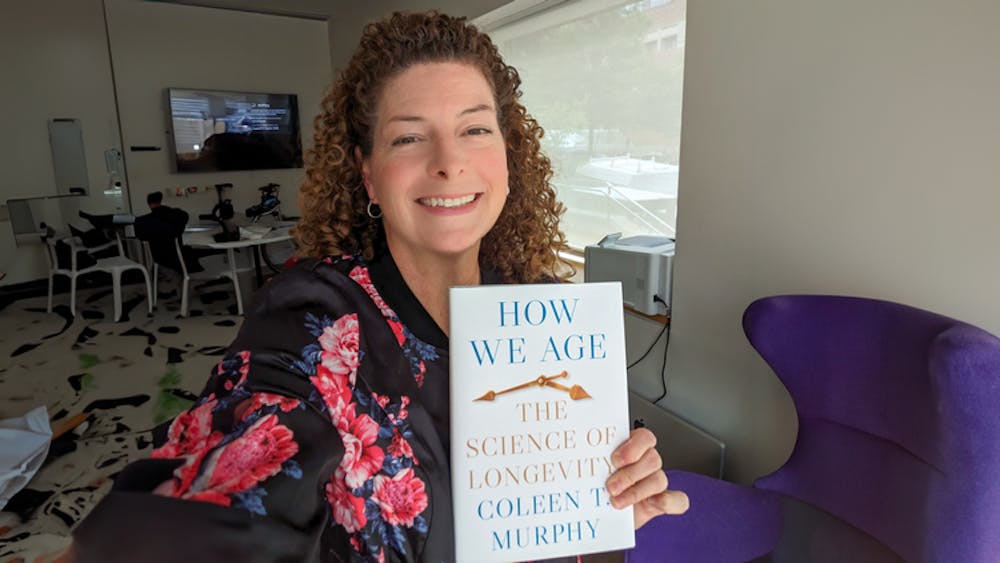 Curly-haired professor smiling and holding up her book that says “How We Age: The Science of Longevity, by Colleen Murphy.”