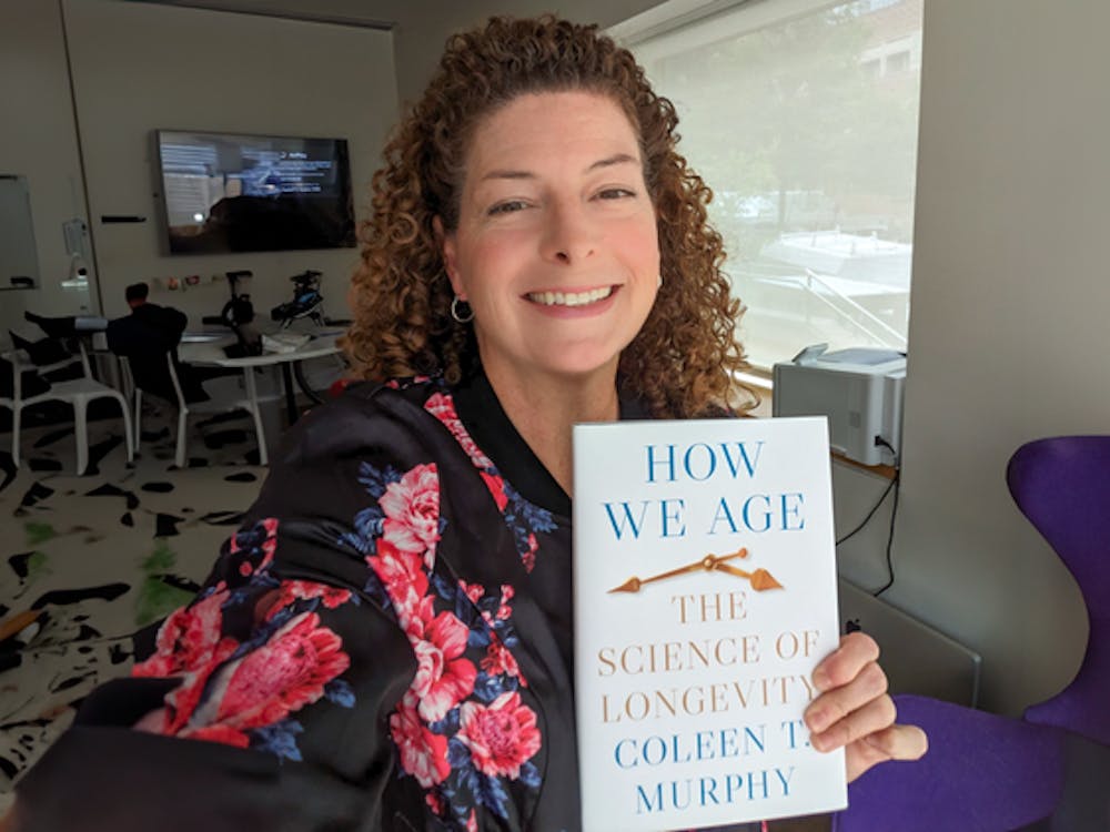 Curly-haired professor smiling and holding up her book that says “How We Age: The Science of Longevity, by Colleen Murphy.”