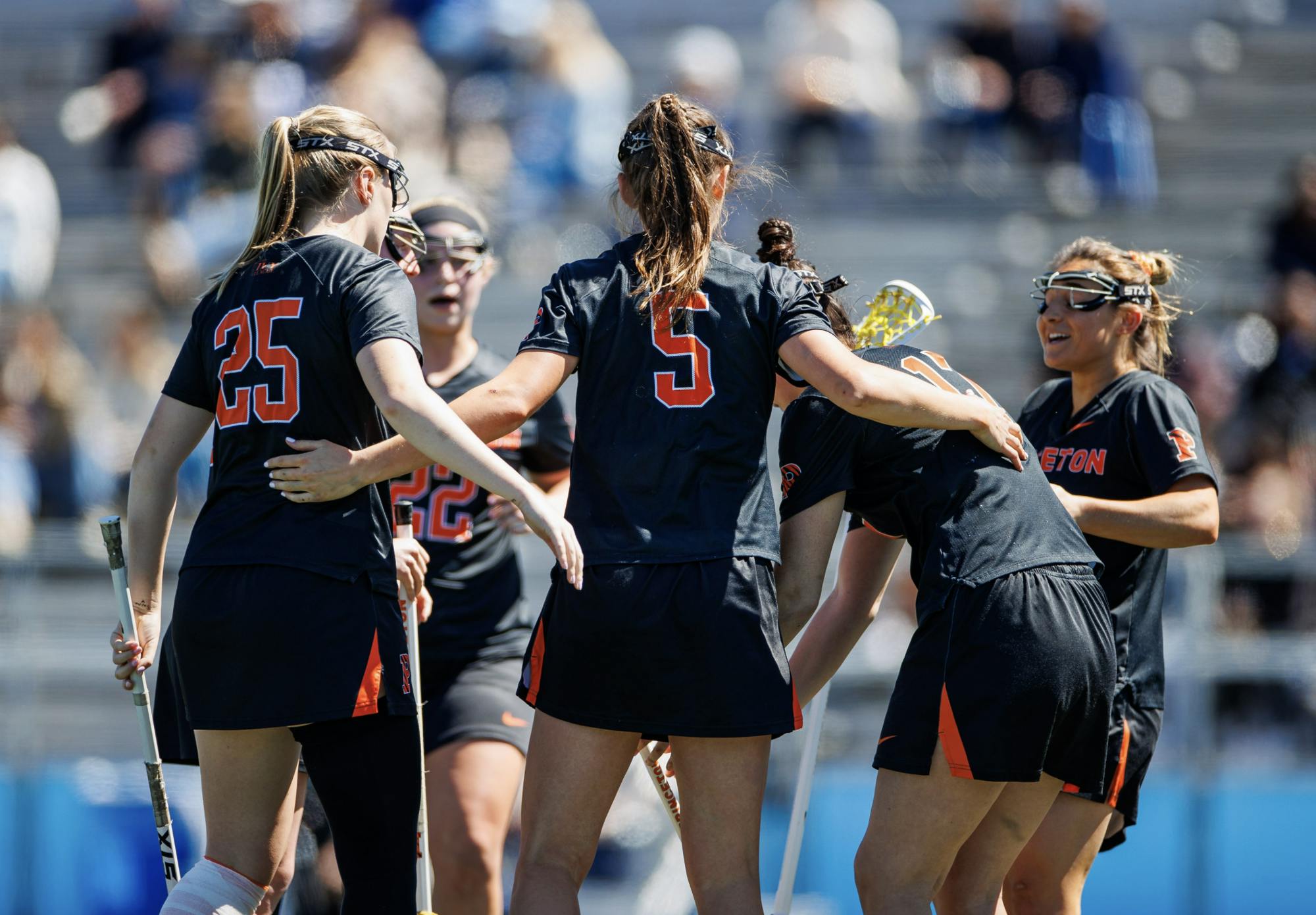 A group of players in a huddle with black jerseys on and holding lacrosse sticks.