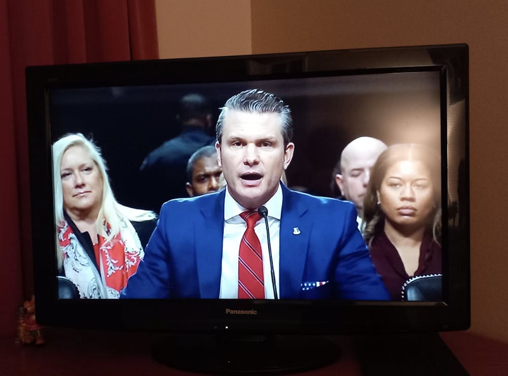 A man wearing a blue blazer and a striped red tie speaks into a microphone on a television screen, with onlookers in the background. 