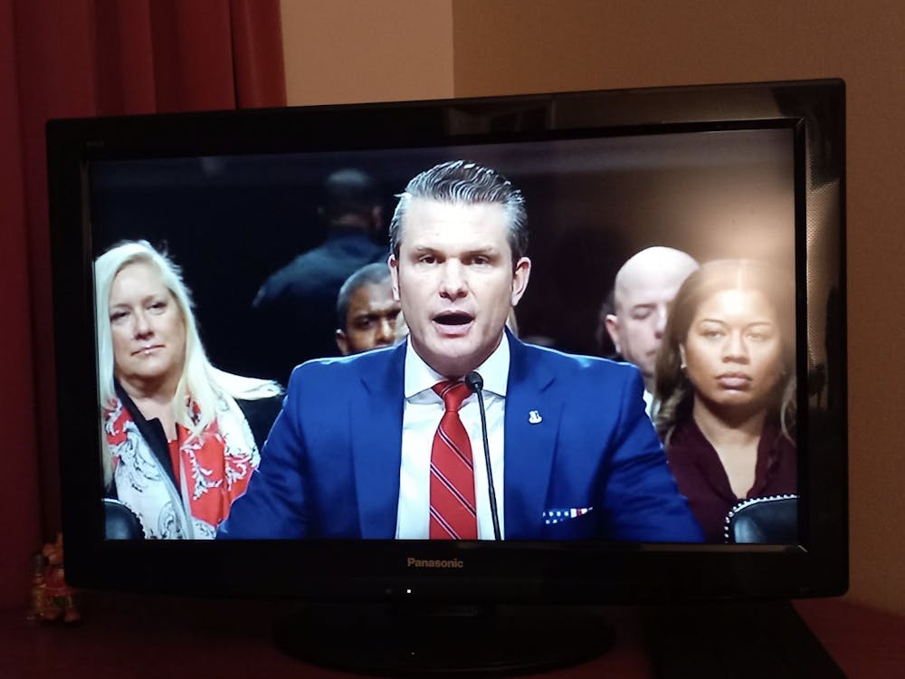 A man wearing a blue blazer and a striped red tie speaks into a microphone on a television screen, with onlookers in the background.