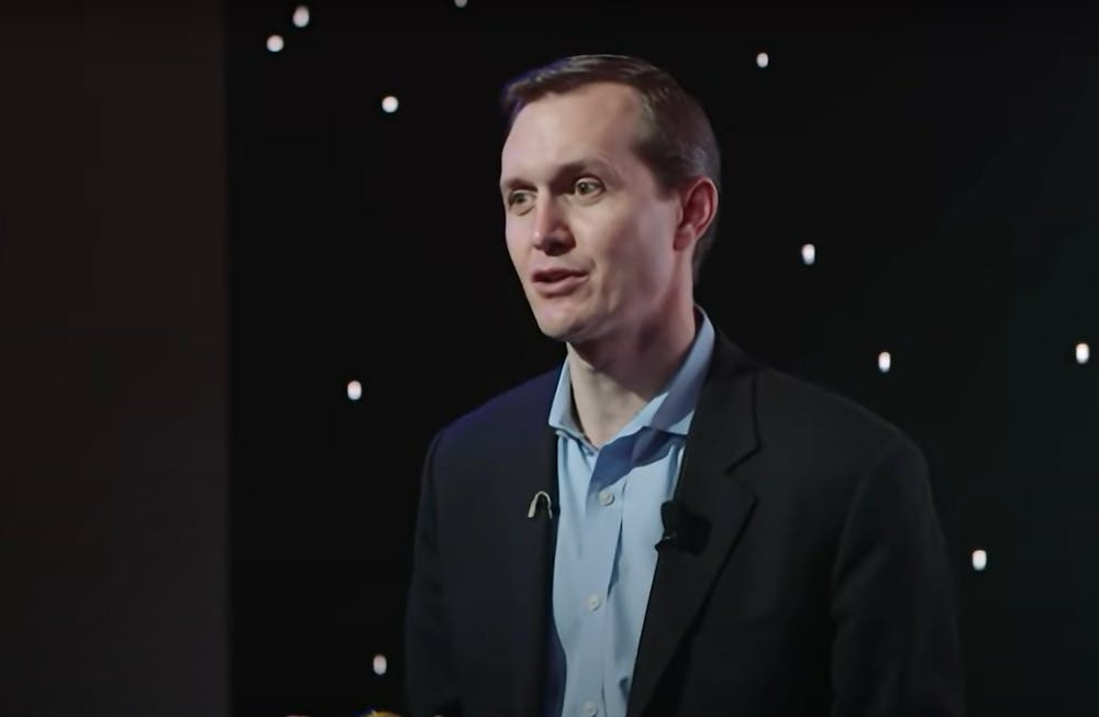 A man in a suit stares to the side in front of a black curtain.