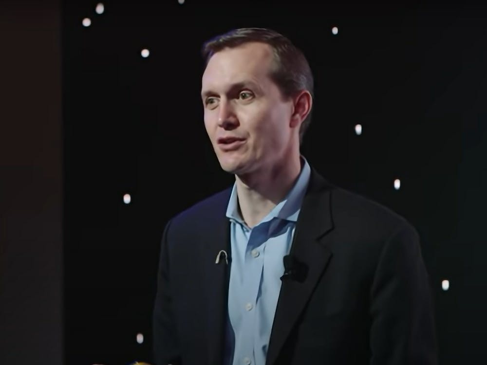 A man in a suit stares to the side in front of a black curtain.