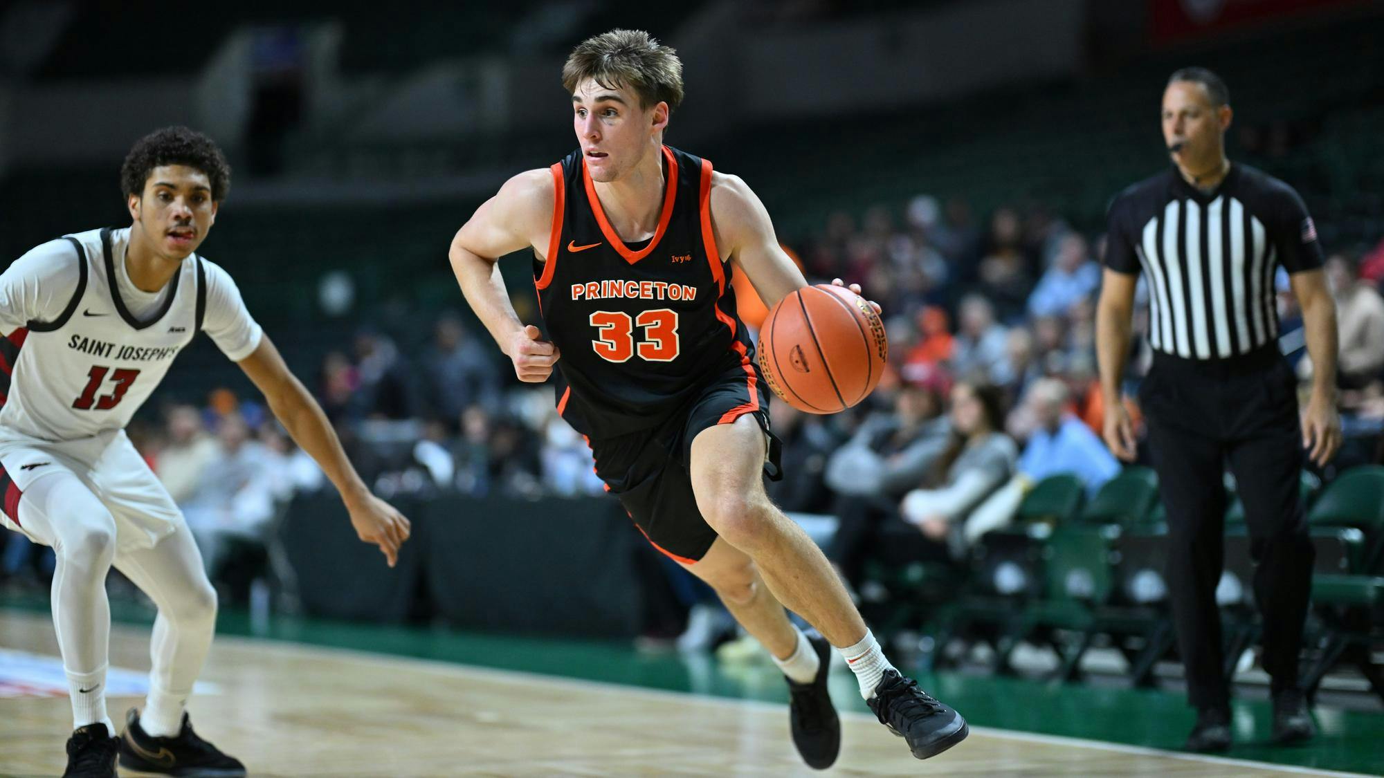A Princeton men’s basketball player drives toward the basket during a game against Saint Joseph’s, dribbling with his right hand as a defender closes in and a referee watches from the baseline.
