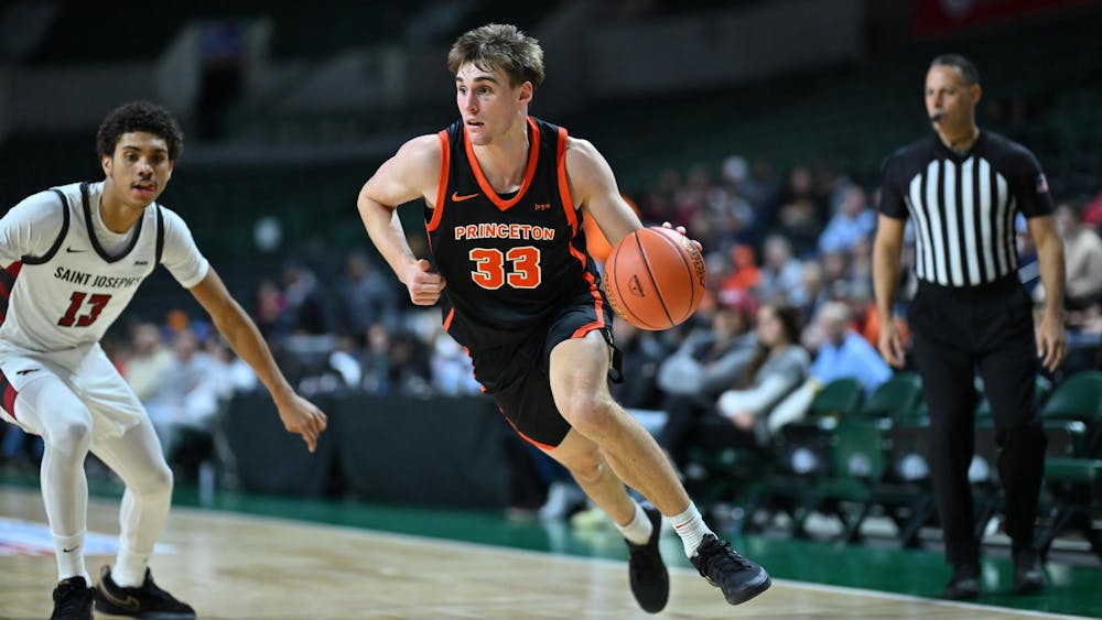 A Princeton men’s basketball player drives toward the basket during a game against Saint Joseph’s, dribbling with his right hand as a defender closes in and a referee watches from the baseline.
