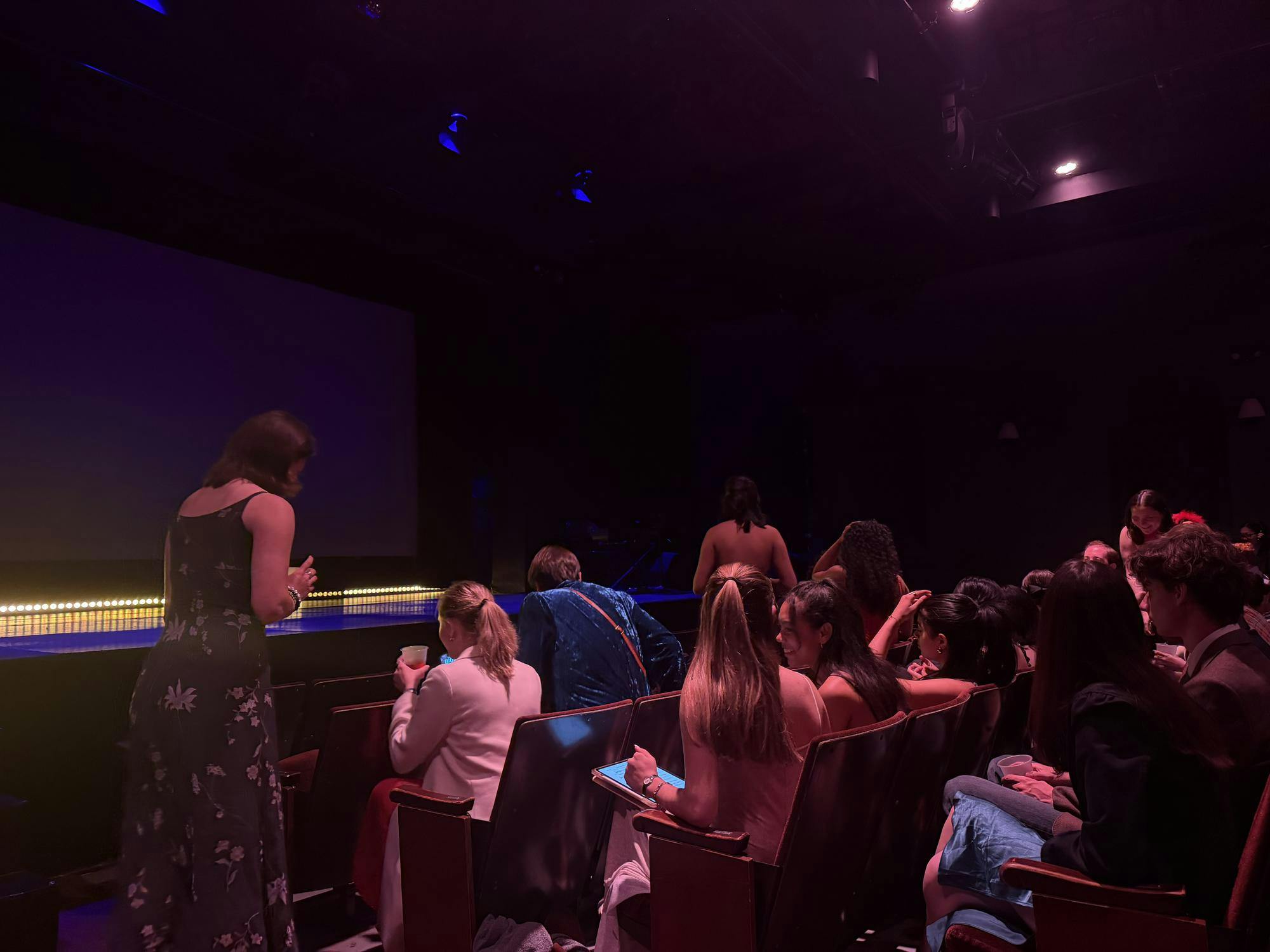 A stage lit with yellow floor lights sits in the back as students dressed up talk amongst themselves. 