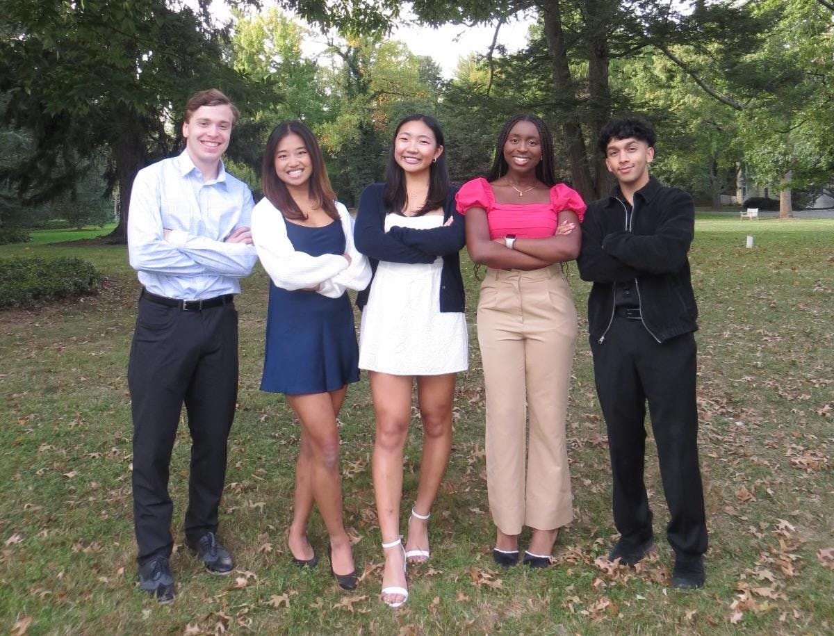 A group of five individuals stand next to each other on a green lawn with trees.