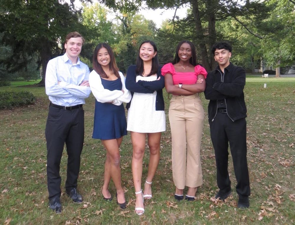 A group of five individuals stand next to each other on a green lawn with trees.