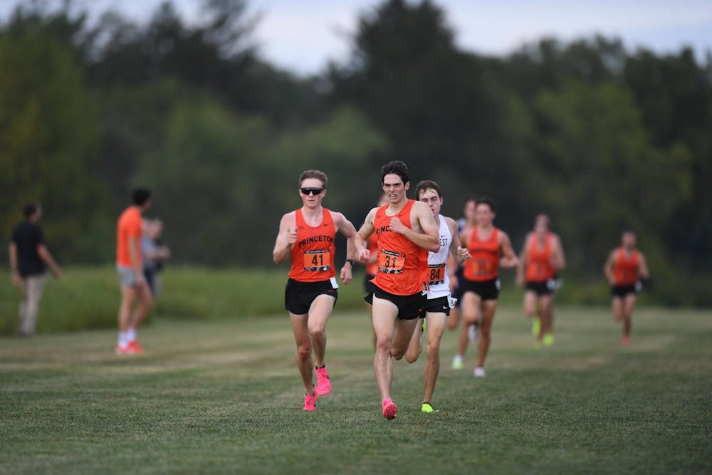 Two runners in orange Princeton uniform trailed by a runner in a white uniform race on green grass. 