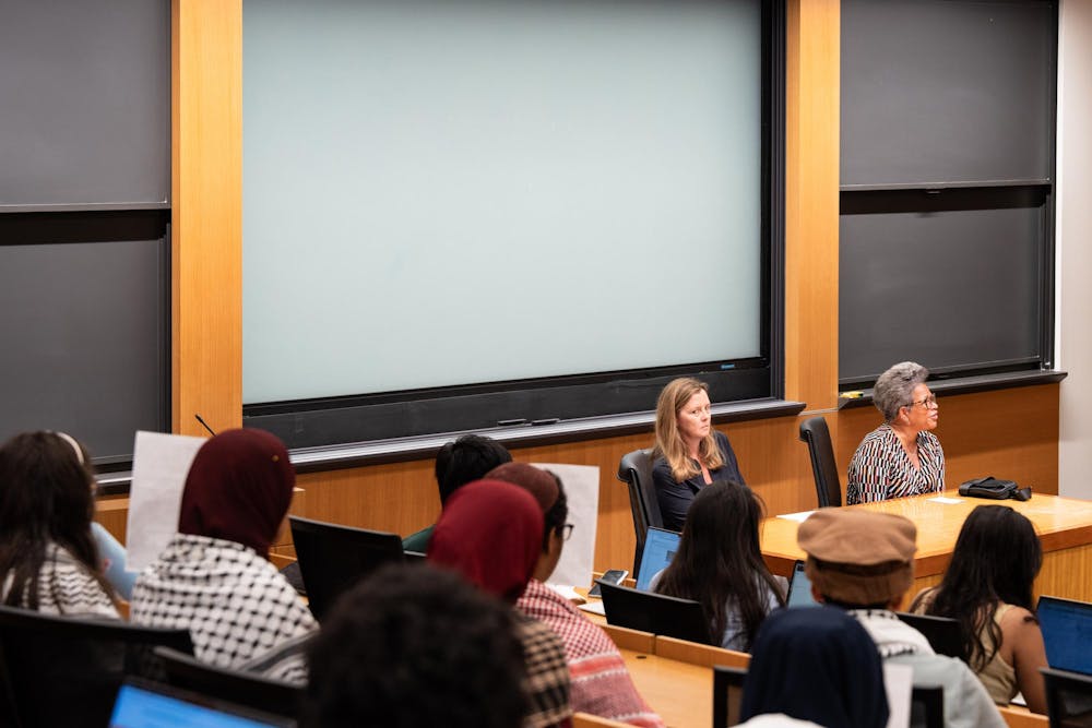 Two women sit at a wooden table, addressing a group of people. Behind the women is a large white screen. Some members of the group hold up posters.