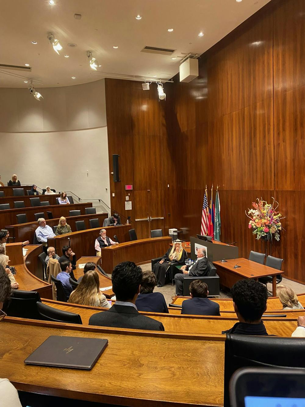 A group of people sit at curved, tiered desks in a wood-paneled room. Two men, one in a keffiyeh, sit in armchairs and speak in the center of the room.