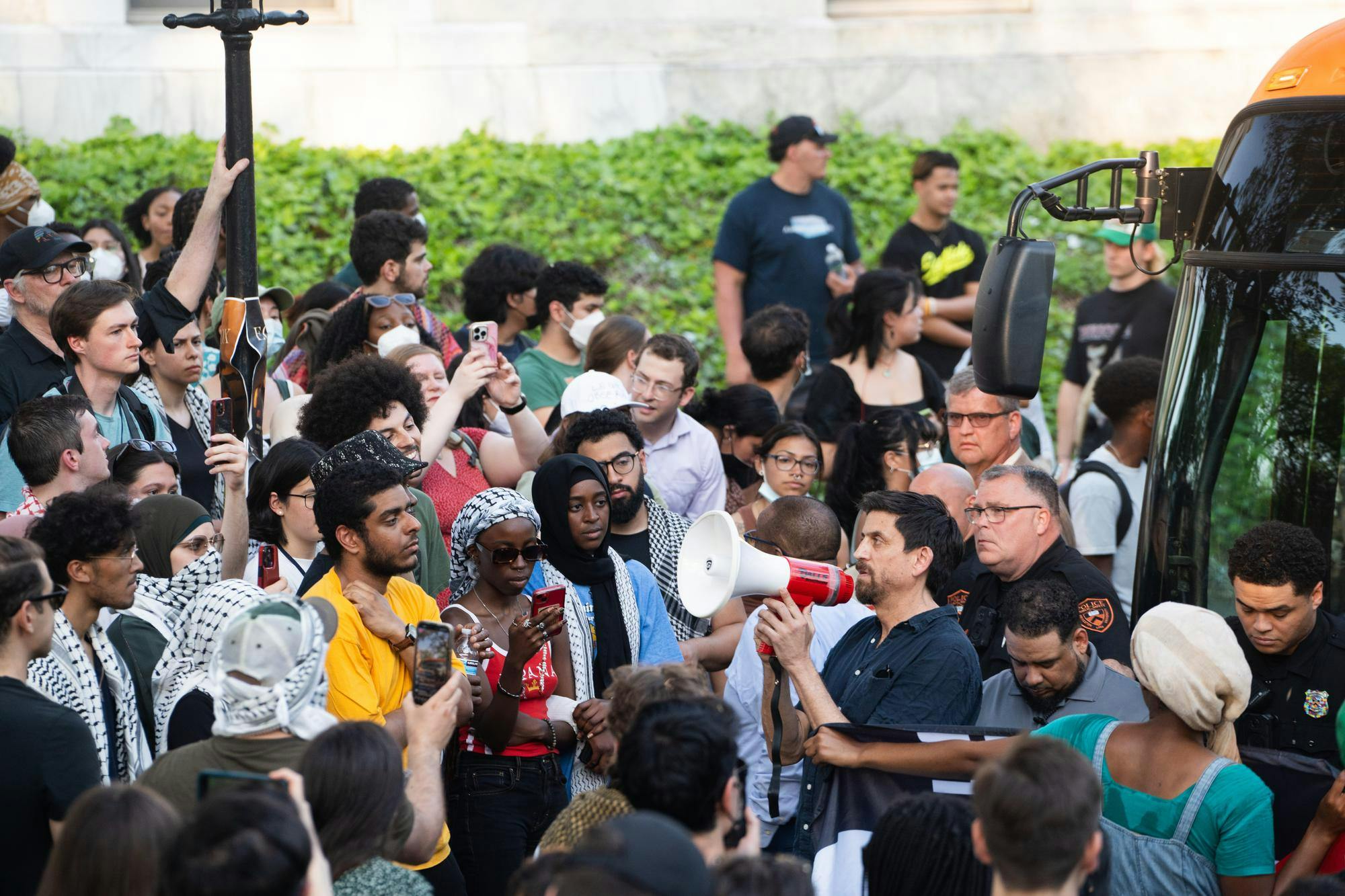 A man holding a megaphone faces towards a crowd in front of a bus. 