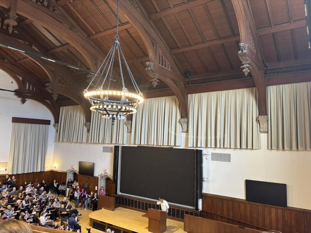 A beige room with a brown, vaulted wood ceiling has a black chandelier hanging from the middle of the ceiling. There are many people sitting watching Everett who is standing on a stage at the front of the room. Everett is wearing a white shirt.