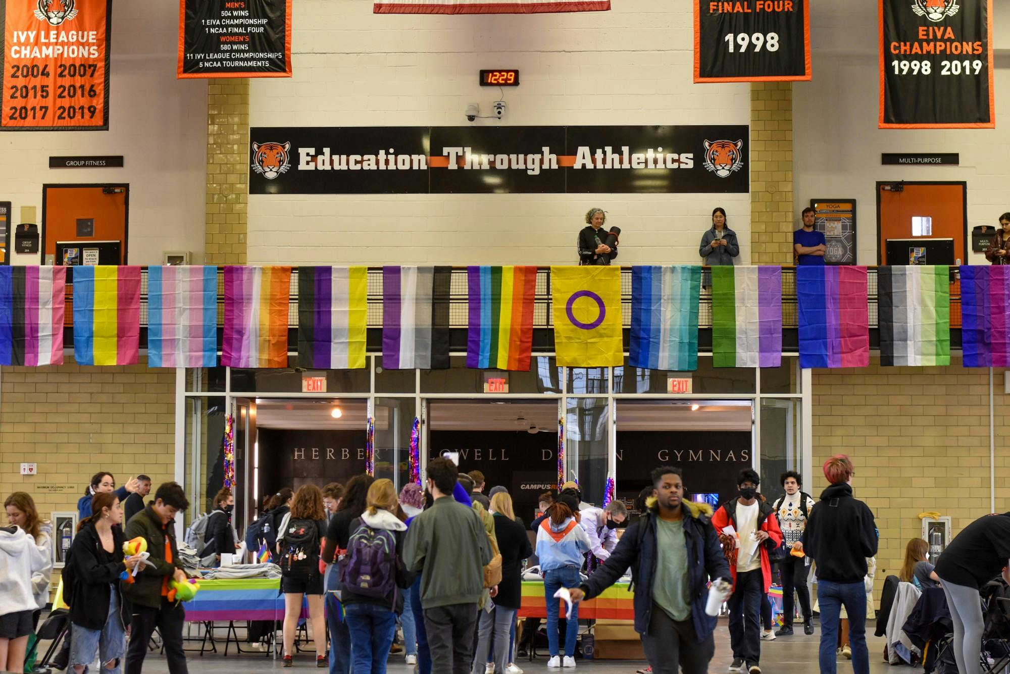 People stand in a gymnasium, with various Pride flags draped over a balcony over the entrance.