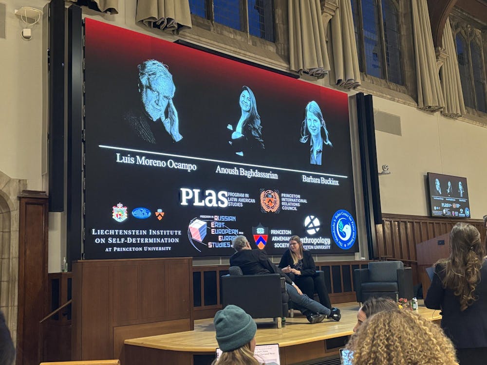 A man and woman sit on a stage in chairs. They sit in front of a screen that shows the name of three panelists under a photo of them. From left to right: "Luis Moreno Ocampo, Anoush Baghdassarian, Barbara Bucking".